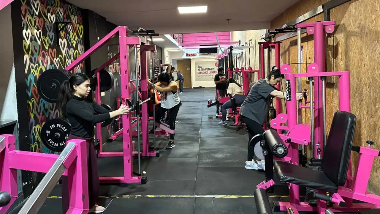 Interior de un gimnasio para mujeres. En el lugar, varias mujeres están entrenando con máquinas de musculación fucsia. A la izquierda hay una pared decorada con un mural de corazones y hay un cartel que dice "Acá no competimos, crecemos juntas"