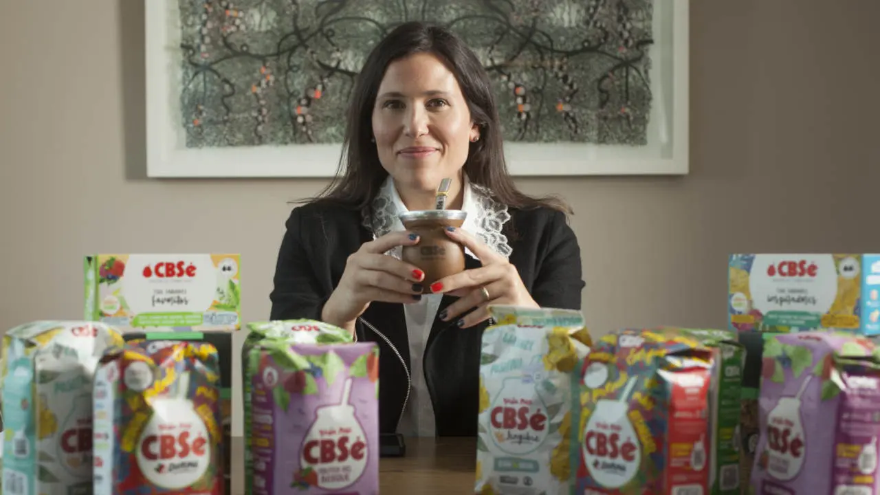 Sol Orquera, CEO de CBSé sentada sostiene un mate y frente a ella, sobre una mesa, hay exhibidos varios paquetes y cajas de productos de yerba mate CBSé