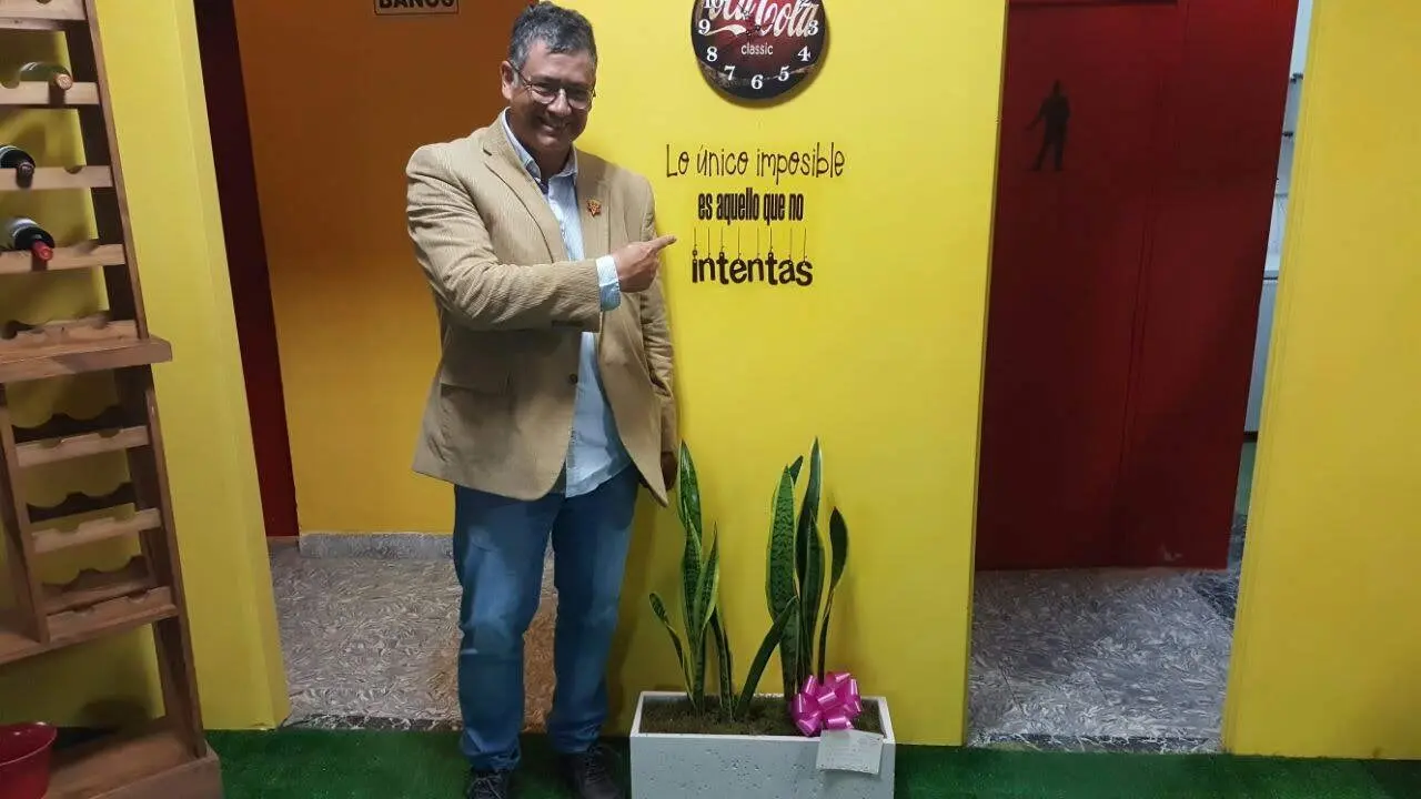 Fabián Sánchez Rojas sonríe con saco beige y jean, señalando en una pared amarilla la frase: "Lo único imposible es aquello que no intentas"   