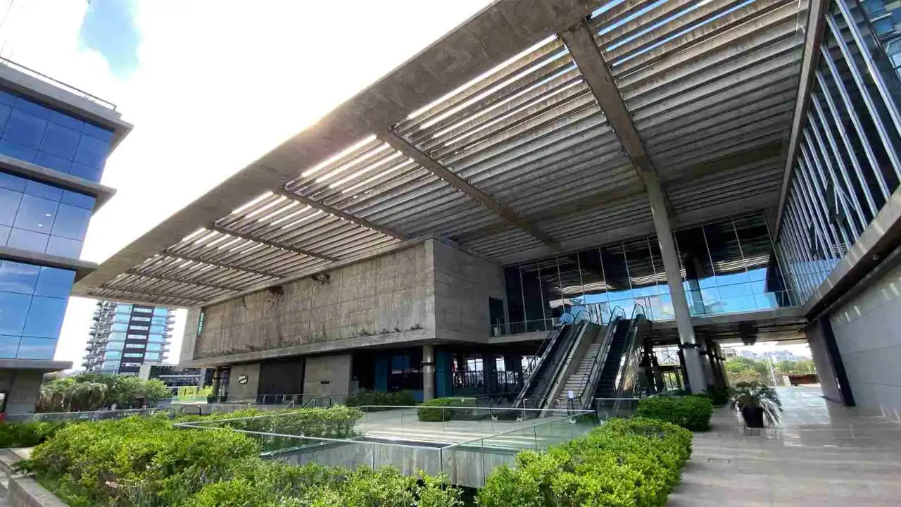Ingreso al edificio Al Rio, con estructuras de hormigón, escaleras mecánicas y vegetación verde en la parte delantera