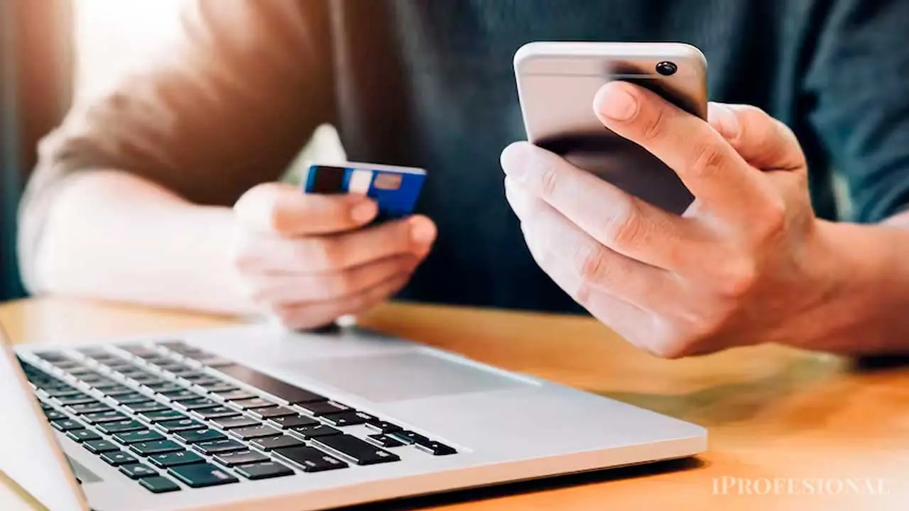 Un hombre con un celular en una mano y una tarjeta de crédito en la otra, y delante una notebook