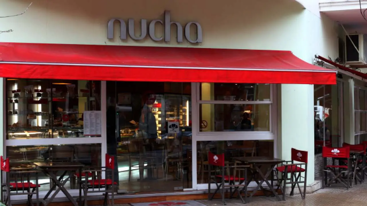 Local de la cafetería Nucha en una esquina con toldo rojo y sillas y mesas en la vereda