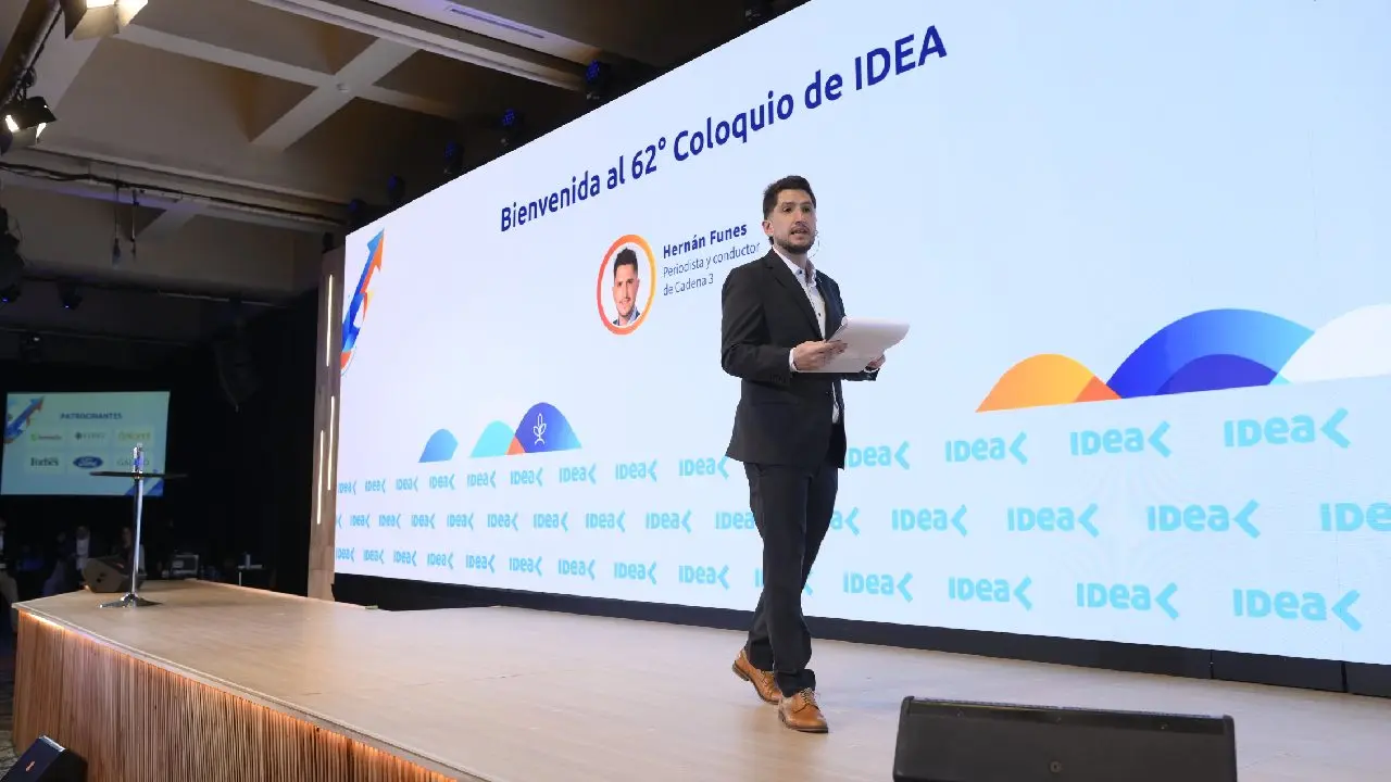 Conductor vestido de traje oscuro hablando arriba de un escenario en el Coloquio de Idea con una pantalla atrás
