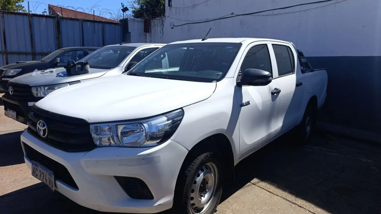 camioneta Toyota Hilux blanca. A su lado hay otras dos camionetas estacionadas en un patio pavimentado con una pared blanca y paredón con alambre de púas al fondo