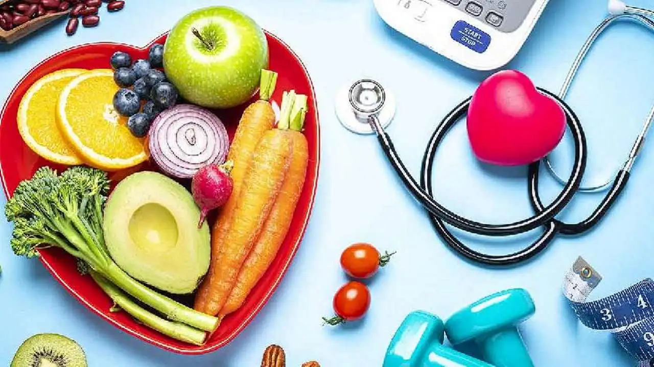 Una fuente con forma de corazón llena de frutas y verduras, al lado unas pesas, una balanza y un estetoscopio con un corazón de juguete