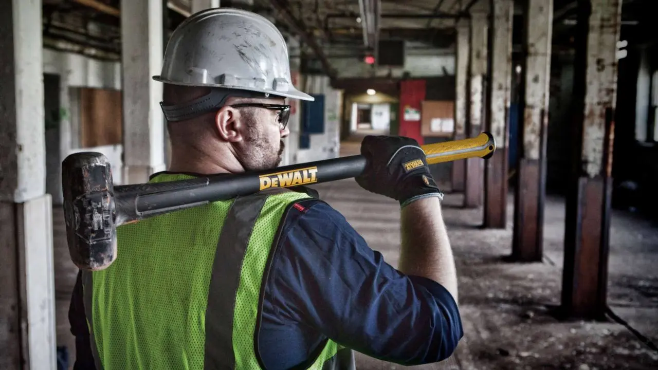 Se ve un trabajador de la construcción con ripa de trabajo y casco. Sosteniene una maza DeWalt sobre sus hombros