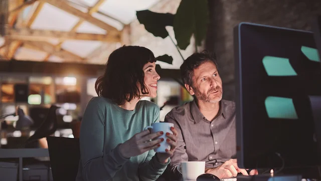 Una mujer con una taza en la mano y un hombre trabajando juntos frente al monitor de una computadora en una oficina