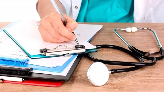 Hombre vestido de médico escribiendo sobre un formulario en una mesa donde hay un estetoscopio. Imagen de archivo.