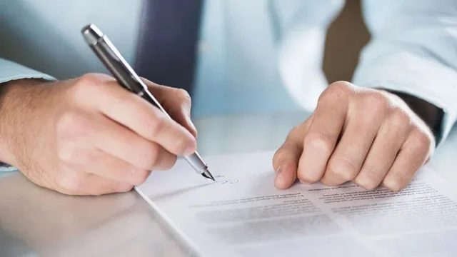 Dos manos de un hombre que en una de ellas tiene una lapicera y está firmando una hoja de papel sobre una mesa. Imagen de archivo