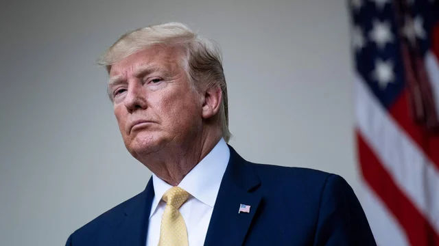 Donald Trump, vestido con traje oscuro y corbata amarilla, posa con mirada desafiante con la bandera de Estados Unidos de fondo