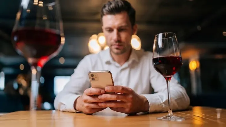 Un hombre con un celular entre sus manos apoyadas en la mesa con dos copas de vino tinto en sus costados. Imagen de archivo