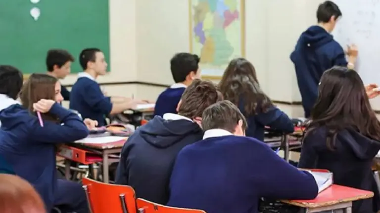 Alumnos y alumnas sentados en sus asientos en el aula mirando a su compañero parado al lado del pizarrón. Imagen de archivo
