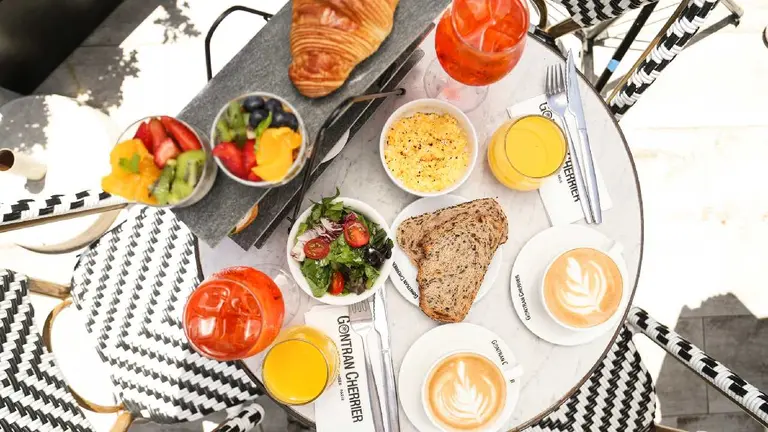 Mesa de un bar al aire libre con un desayuno que incluye cafe, huevo, frutas, juegos y panficados, entre otras cosas