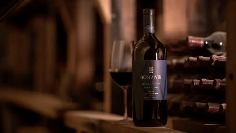 Una botella de vino junto a una copa con vino, detrás hay una bodega con varias botellas recostadas