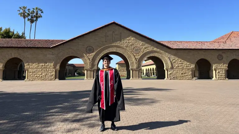 Armando Borda vestido con la toga y el birrete de graduación de Stanford, parado en el centro de un patio del campus.