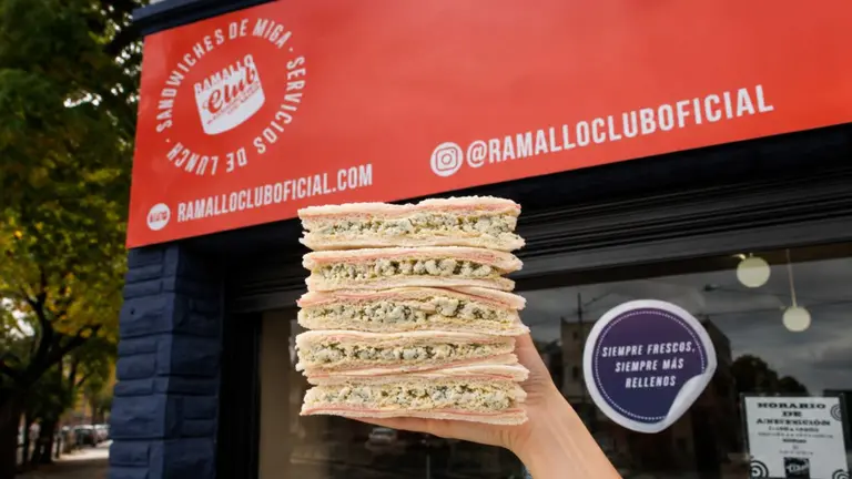 Frente de un local Ramallo Club con una mano llena de sándwiches de miga asomando. Gentileza Ramallo Club
