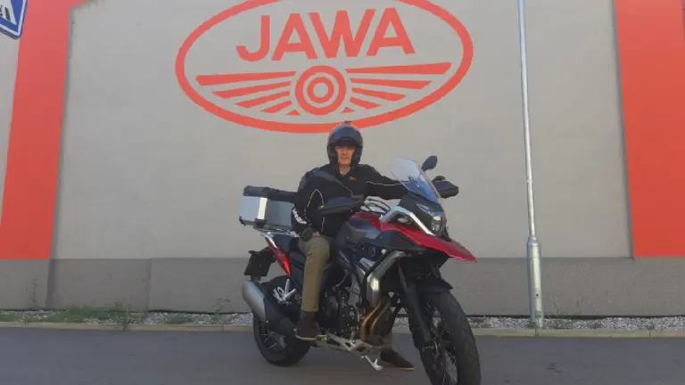 Hombre arriba de una moto de alta cilindrada con un cartel atrás que dice JAWA en letras rojas sobre una pared gris