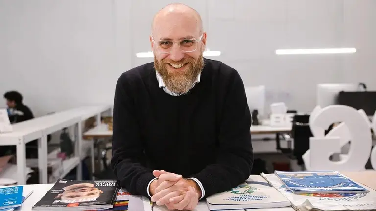 Gonzalo Otalora sonriendo con las manos cruzadas frente a un escritorio con revistas dispersas en el mismo