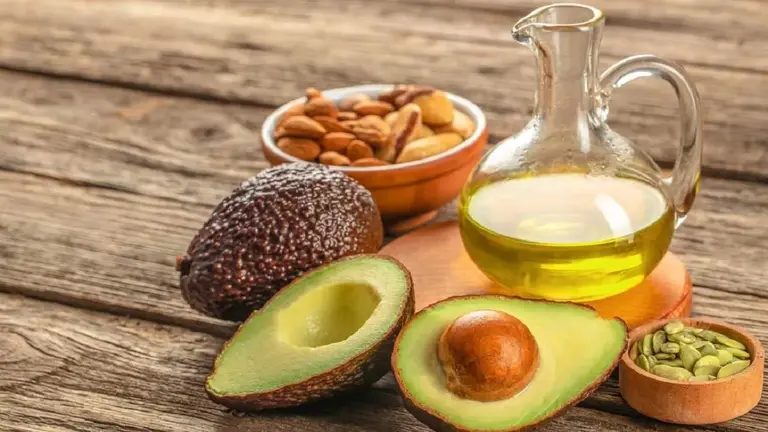 En una mesa, una palta cortada a la mitad y otra entersa, aceite en una botella de vidrio y frutos secos en un plato chico