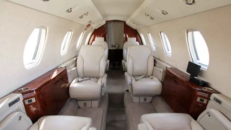 Interior de un avión privado con asientos de cuero, mesas laterales de madera y ventanillas a ambos lados.