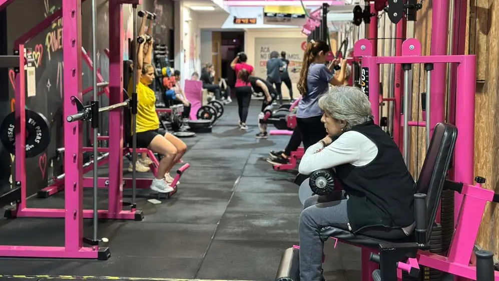 Gimnasio femenino activo con máquinas rosas, pesas y mujeres de distintas edades entrenando