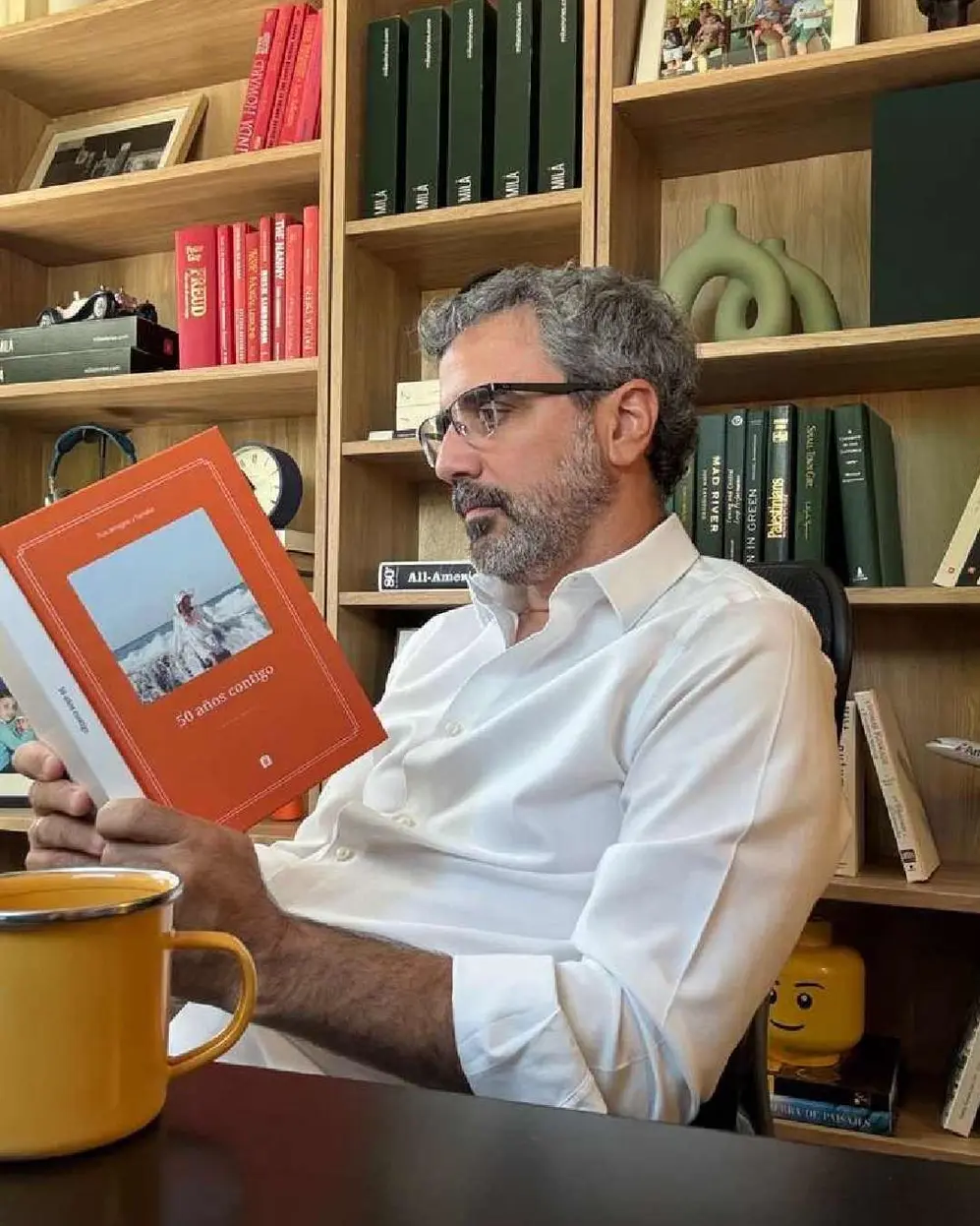 Persona sentada leyendo un libro de memorias con una biblioteca atrás y una taza amarilla adelante