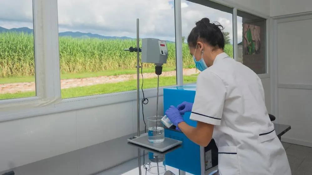Cientifica de espalda con barbijo en labotario haciendo pruebas con una ventana con vista al campo