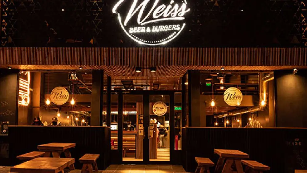 Frente del local de la hambuguersía Weiss de noche con mesas de madera en su parte delantera