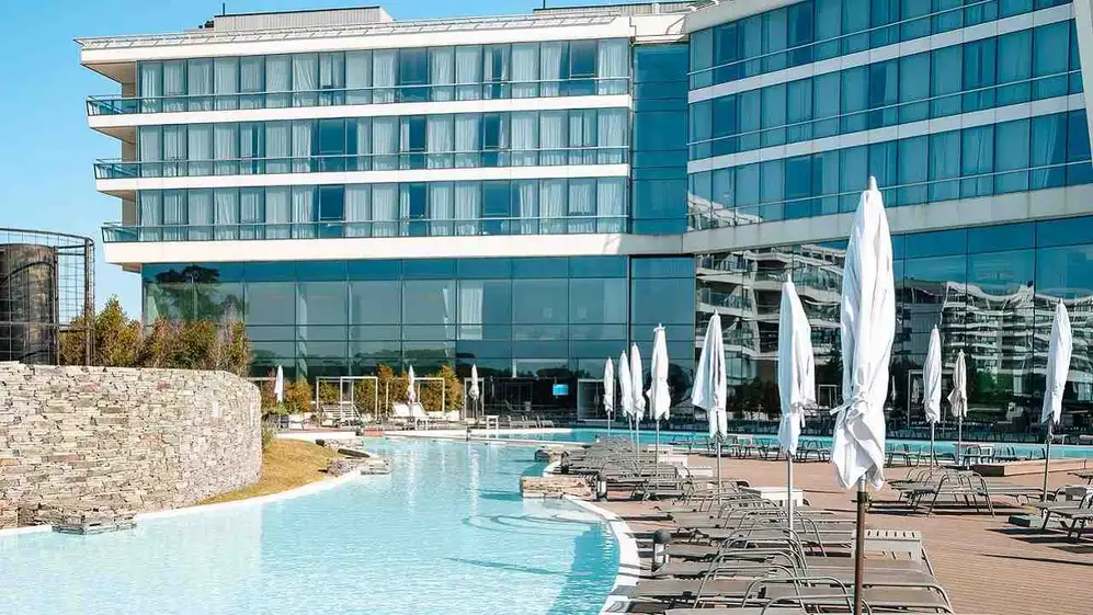 Una piscina con reposeras y sombrillas alrededor y de fondo el hotel Hilton Pilar