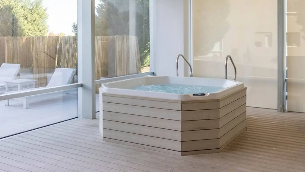 Un spa en una habitación con vidrios y detrás unas reposeras al aire libre