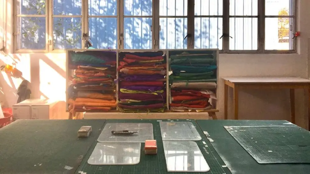 Mesa y objetos de trabajo con un mueble con telas de colores atrás y una ventana