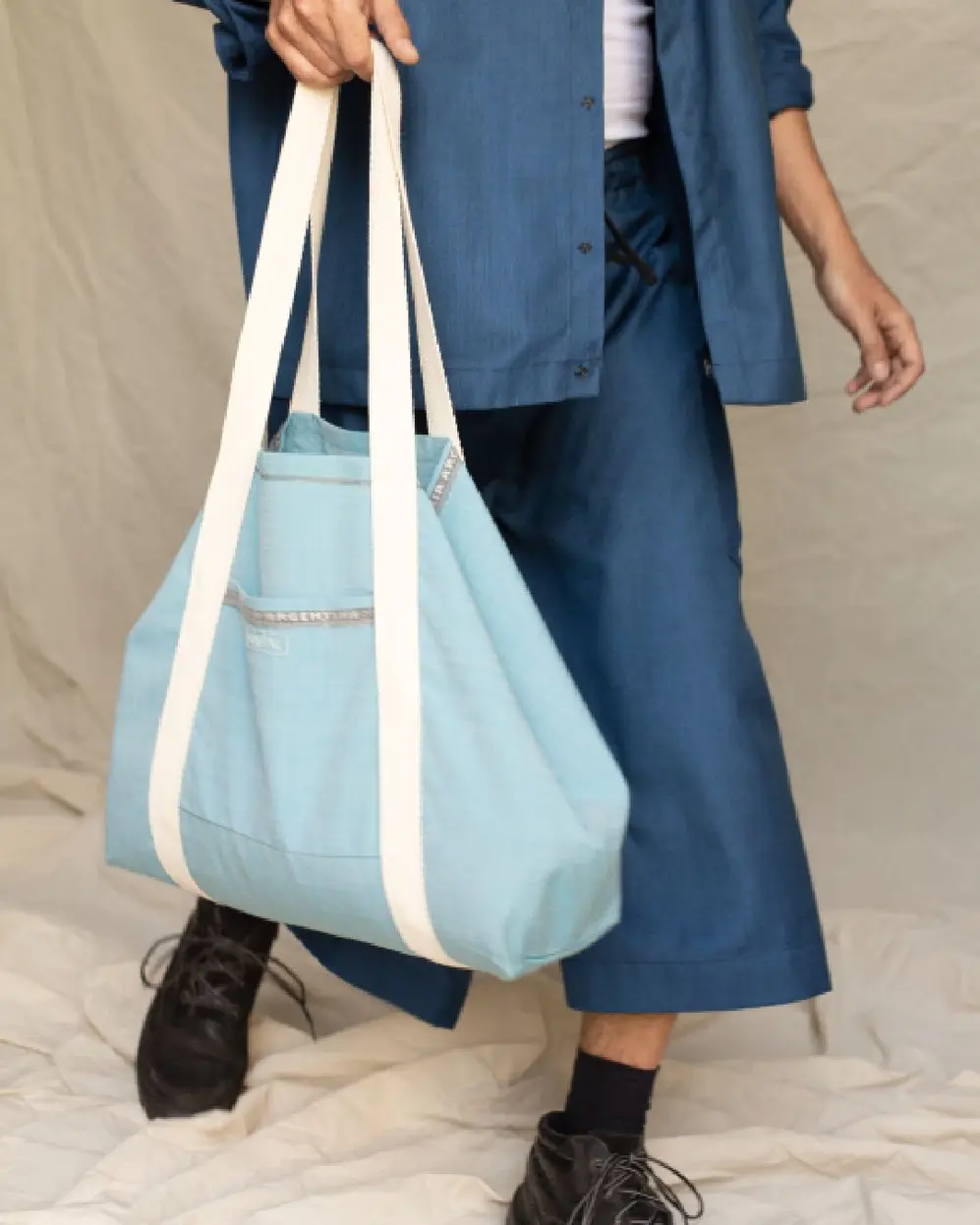 Una persona vestida de azul sosteniendo un bolso de tela color celeste con manijas blancas
