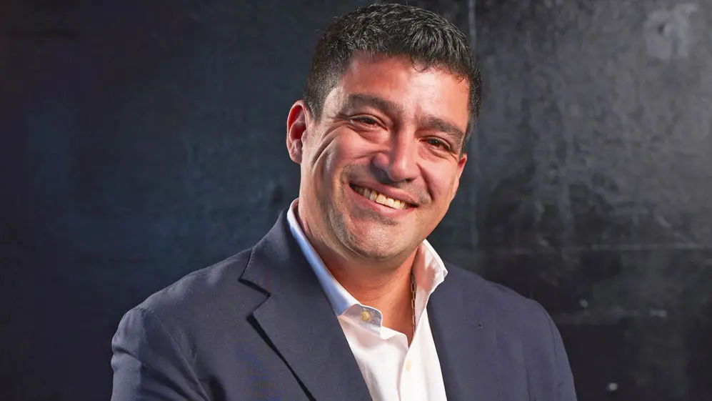 Retrato de Julio Gauna sonriendo, con saco oscuro y camisa blanca, sobre un fondo negro