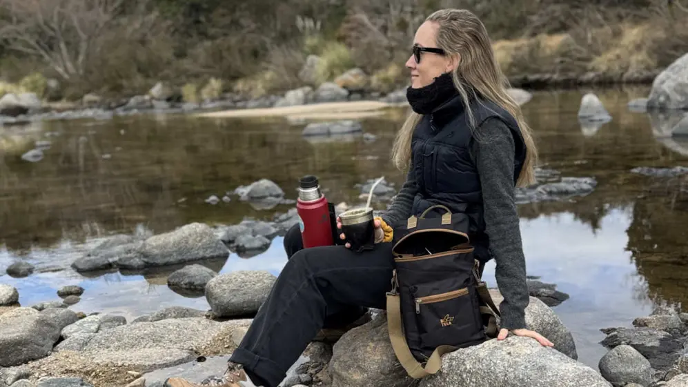 Paula Madero sentada junto al río tomando mate con gafas de sol, termo y un bolso matero de Botela